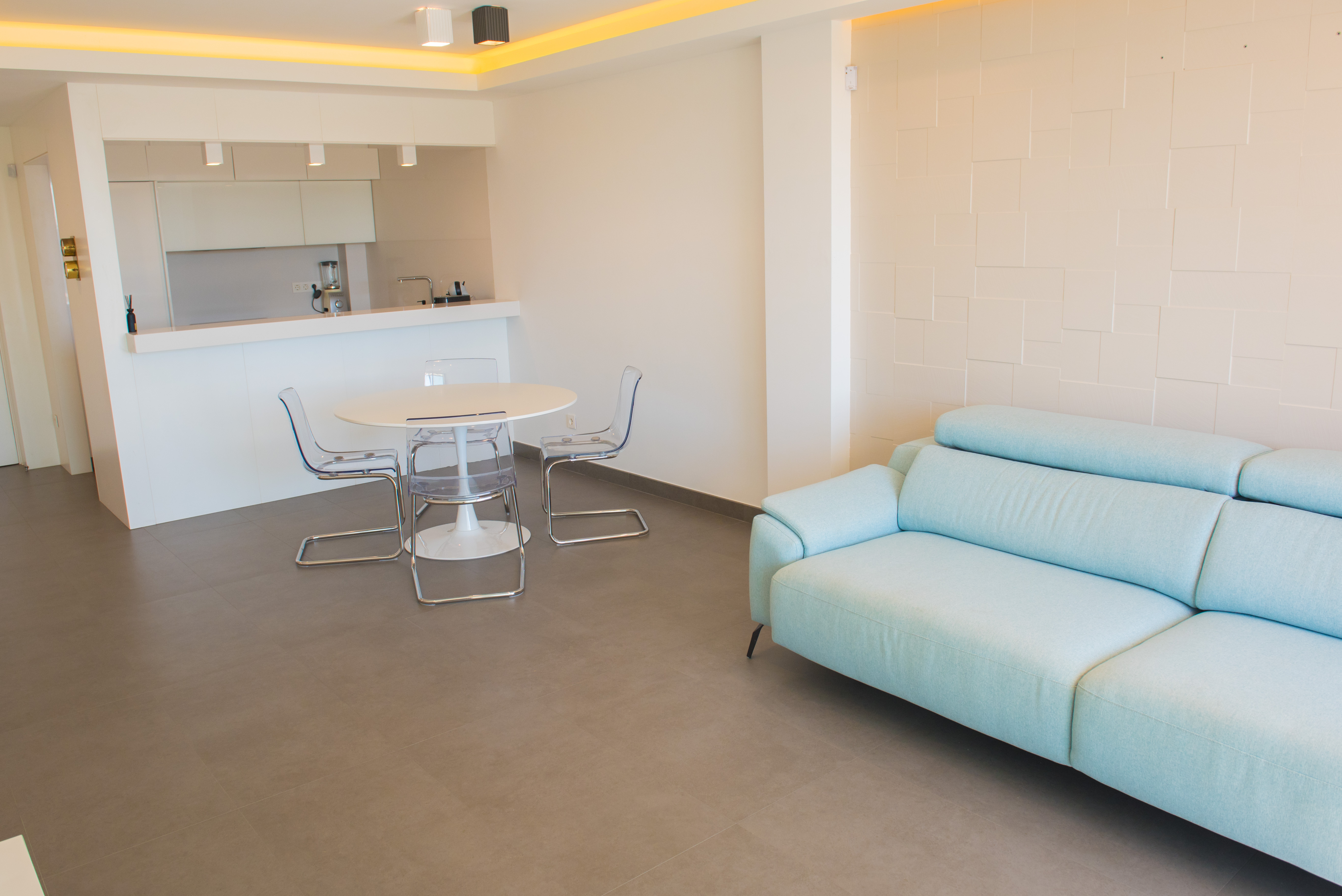 Open-plan living area featuring a light blue sofa, transparent acrylic chairs, and a white dining table. Includes a textured geometric wall and recessed ceiling lighting.