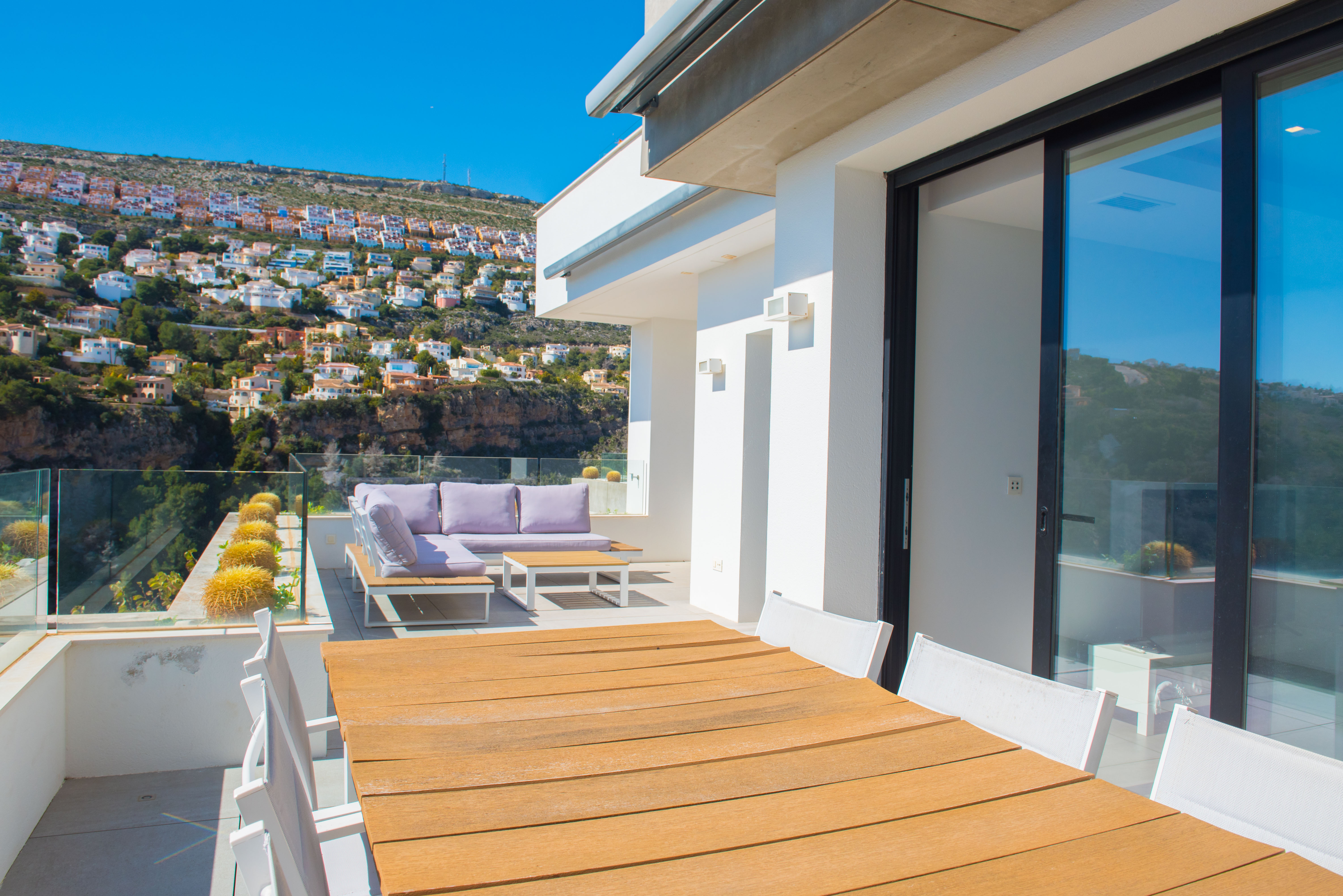 Outdoor terrace featuring a wooden dining table, lilac lounge seating, and glass railings overlooking the hillside urbanization and clear blue sky.