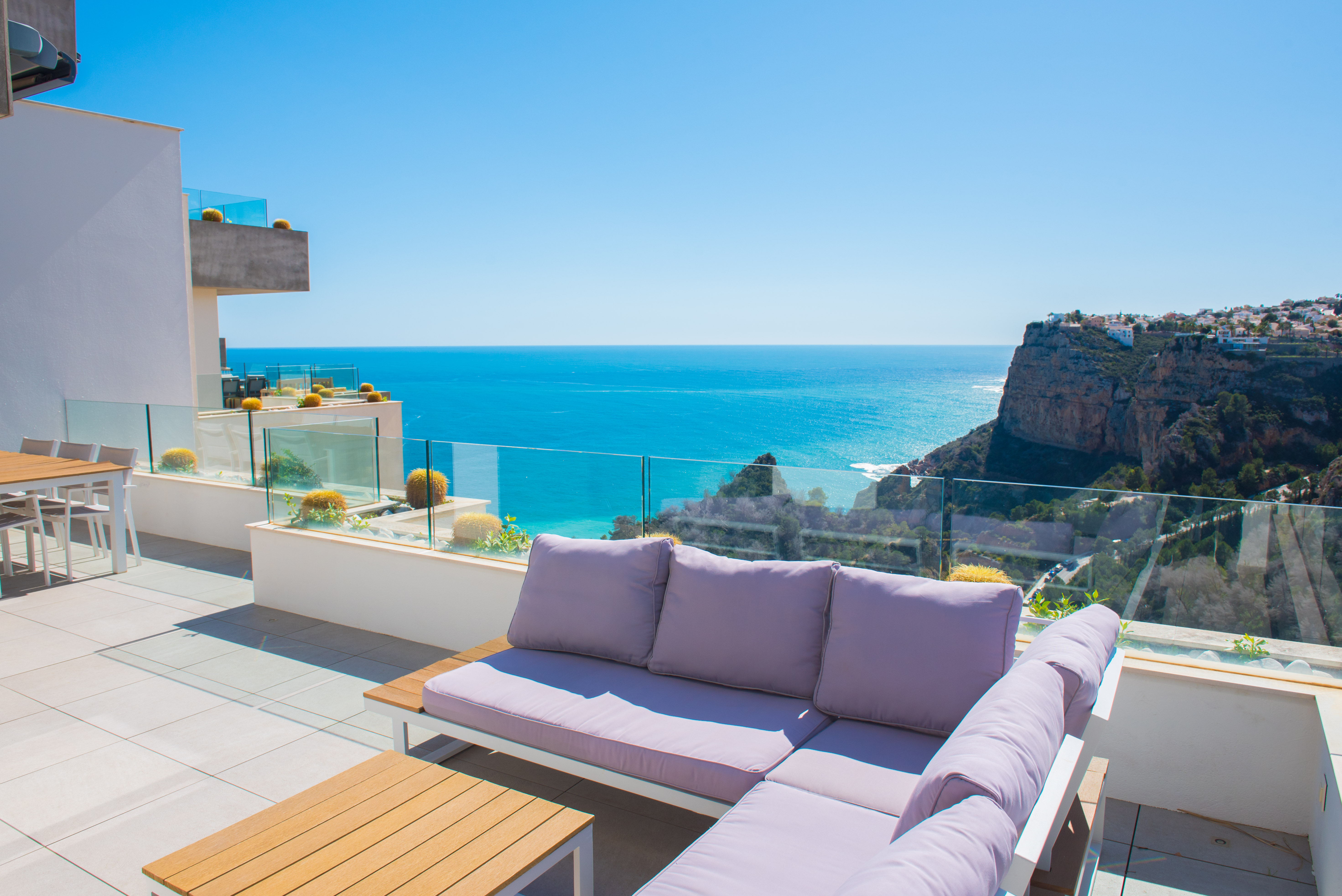 Private terrace featuring an L-shaped outdoor sofa, glass railings, and panoramic views of the Mediterranean Sea and coastal cliffs.
