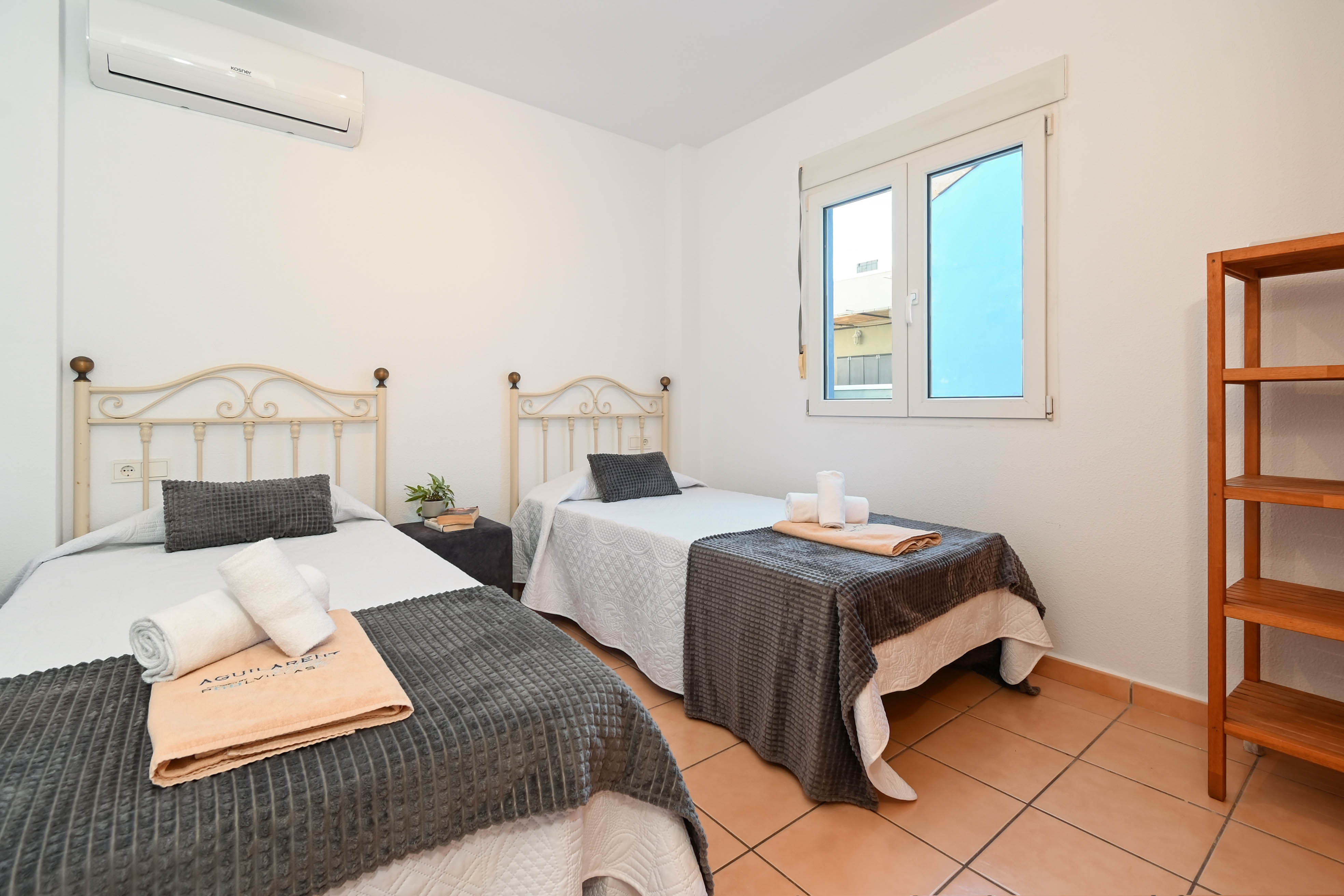 Bedroom featuring twin beds with wrought-iron headboards, terracotta tile flooring, and split air conditioning. Includes a wooden shelving unit and natural light.