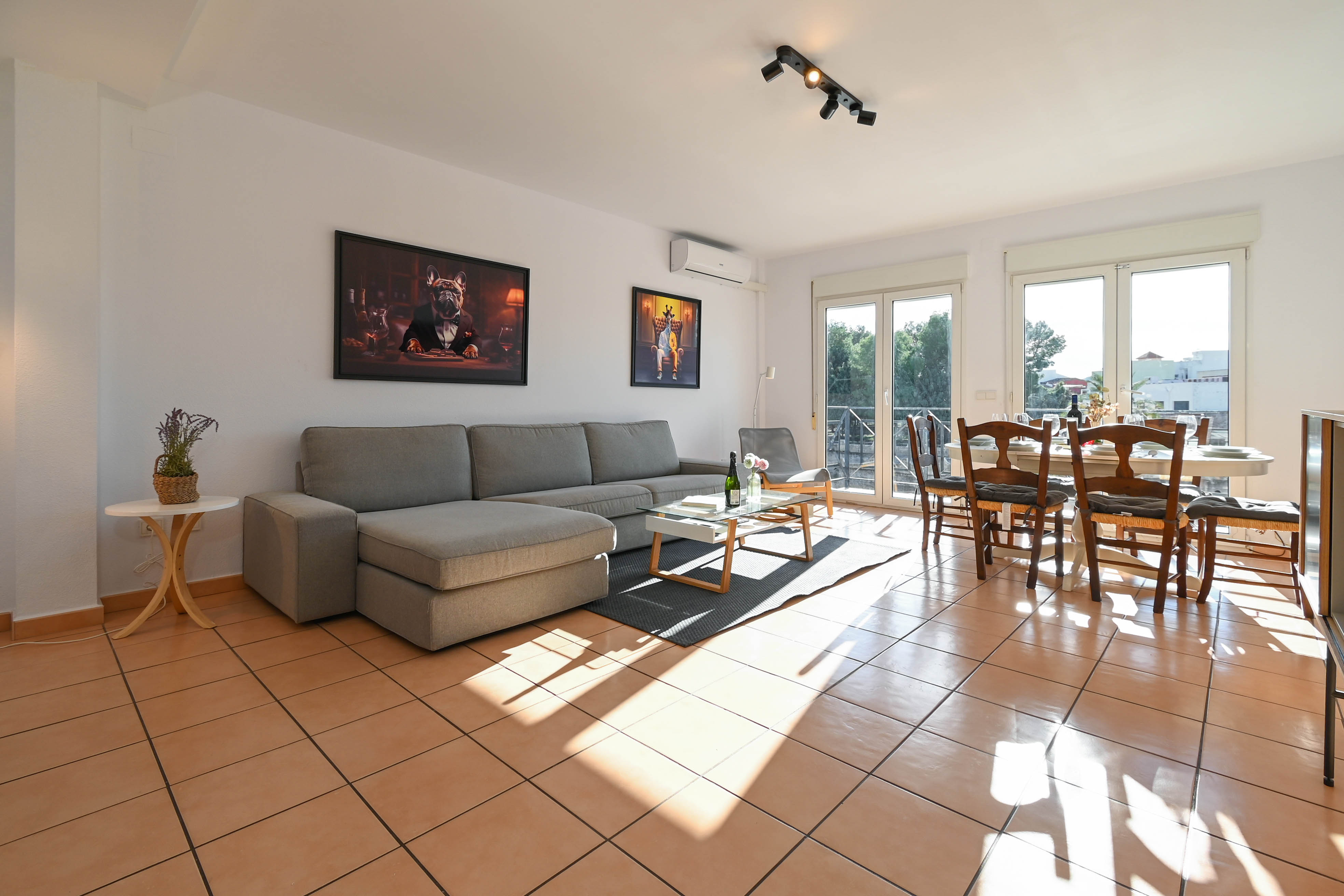 Bright living and dining area featuring terracotta tile flooring, a grey sectional sofa, and glass doors opening to a sunlit balcony.
