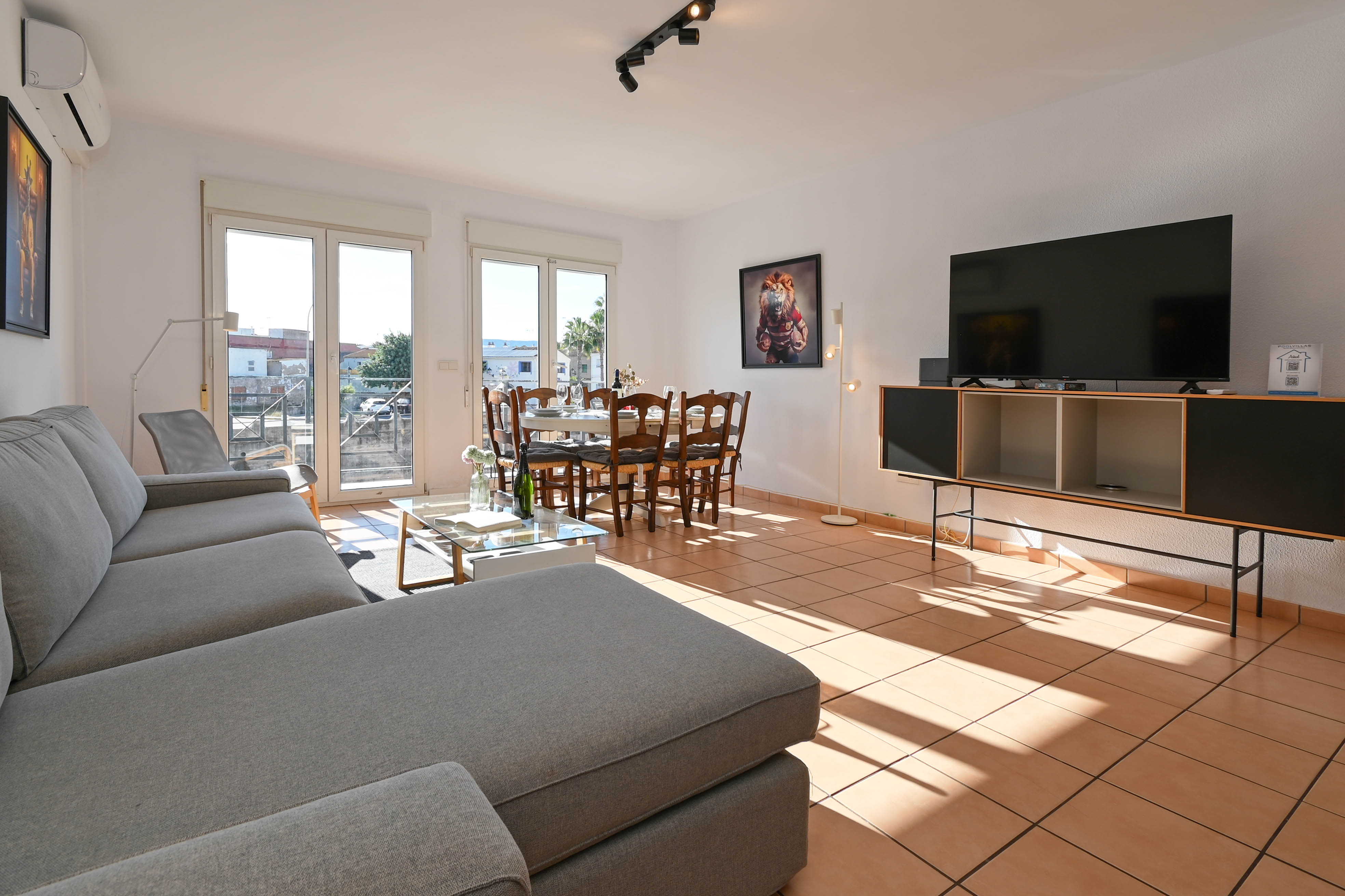 Bright open-plan living and dining area featuring terracotta tile flooring, a large grey sectional sofa, and direct access to the balcony via glass doors.