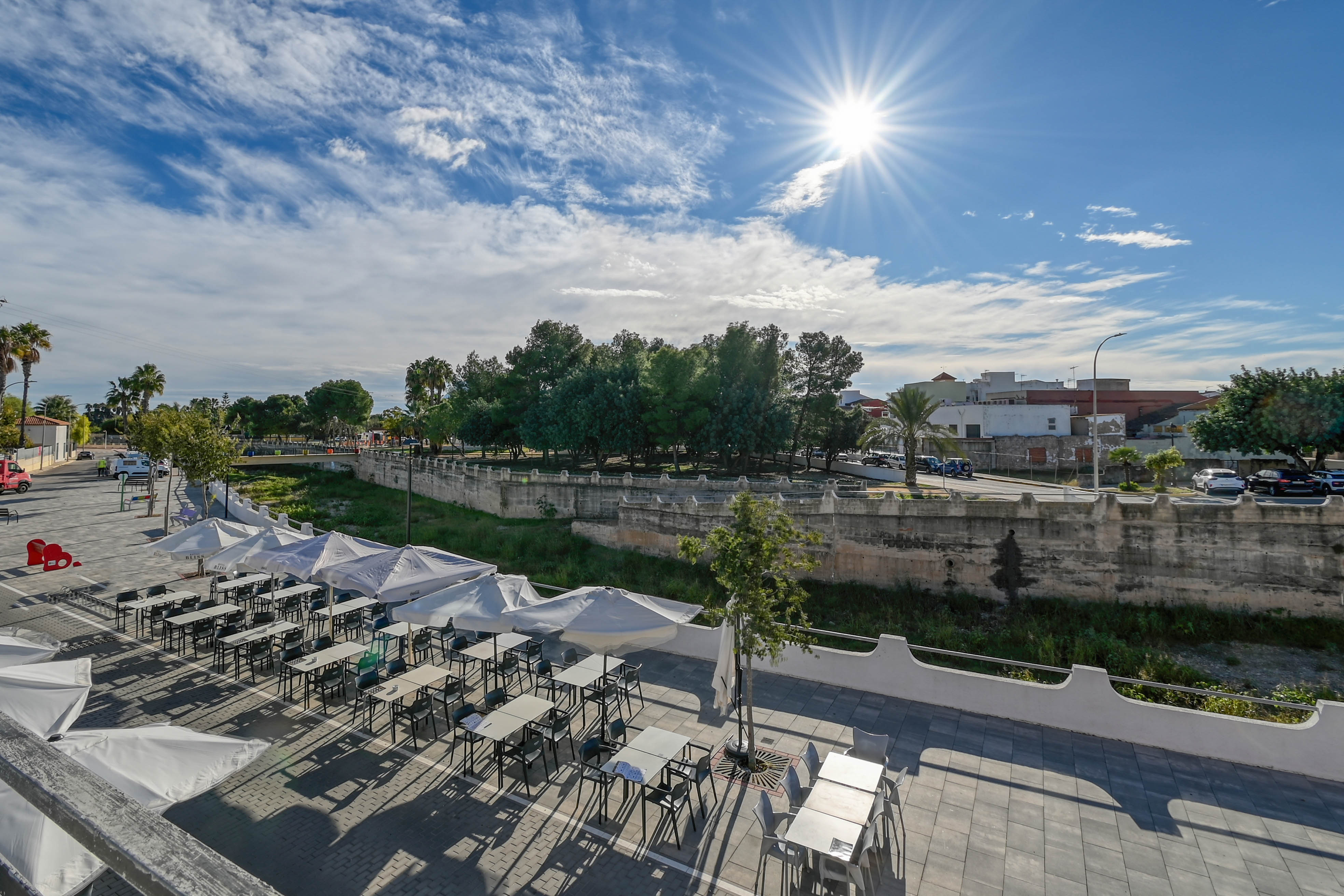 Elevated view of an outdoor terrace featuring white parasols and seating, overlooking a stone-walled canal and lush green trees under a bright sun.