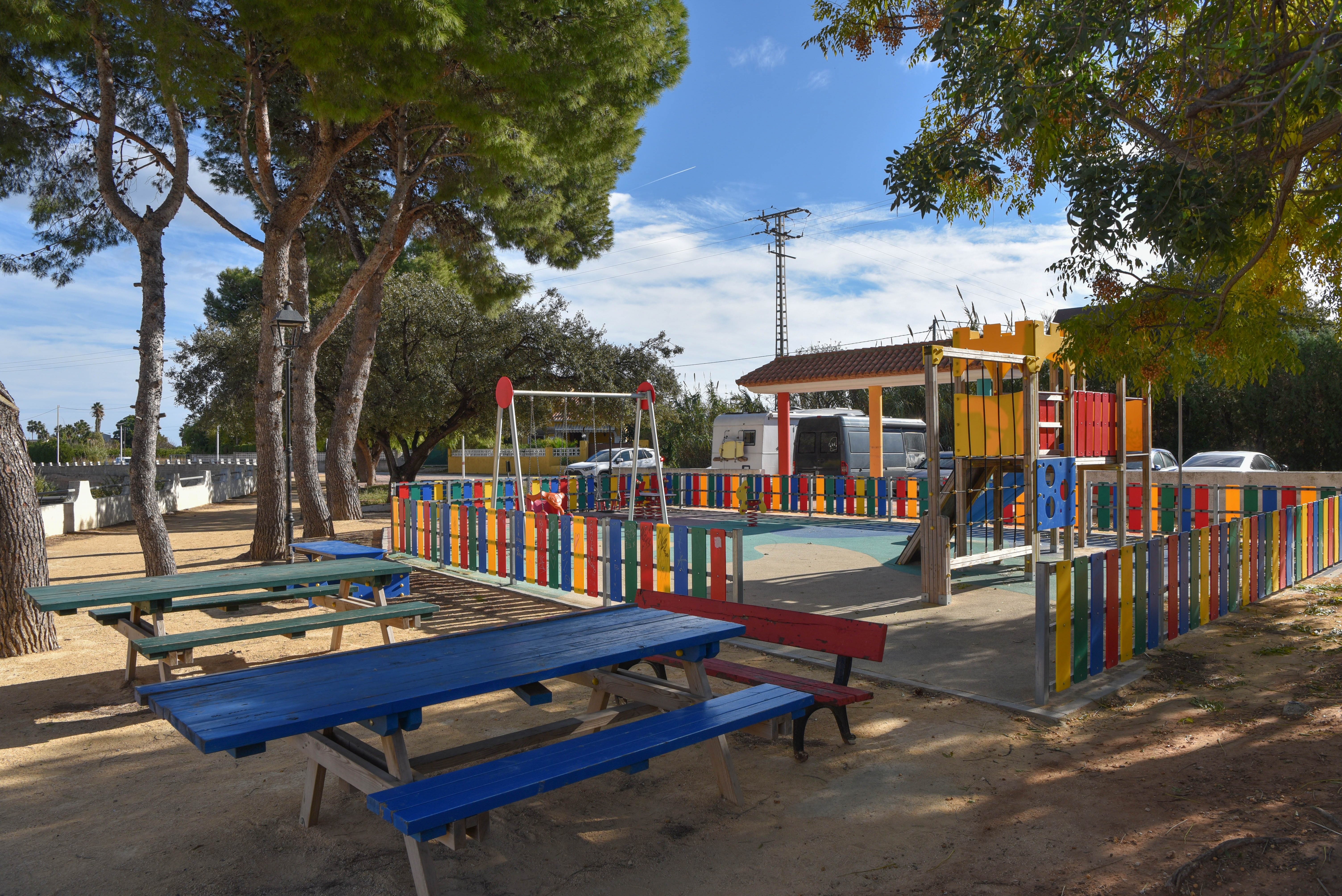 Fenced children's playground featuring colorful play structures and wooden picnic tables shaded by mature pine trees in a sunny outdoor setting.