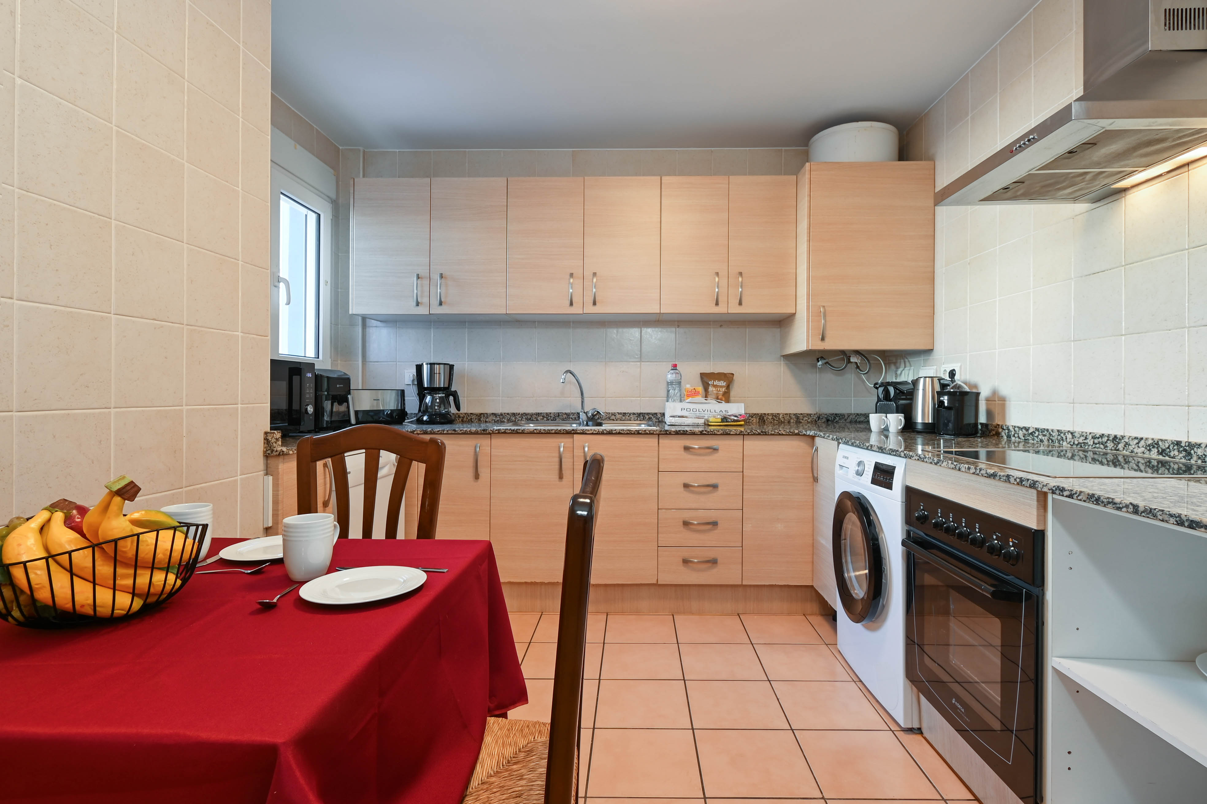 Fully equipped kitchen featuring light wood cabinetry, granite countertops, integrated appliances, and a dining area with a red tablecloth.
