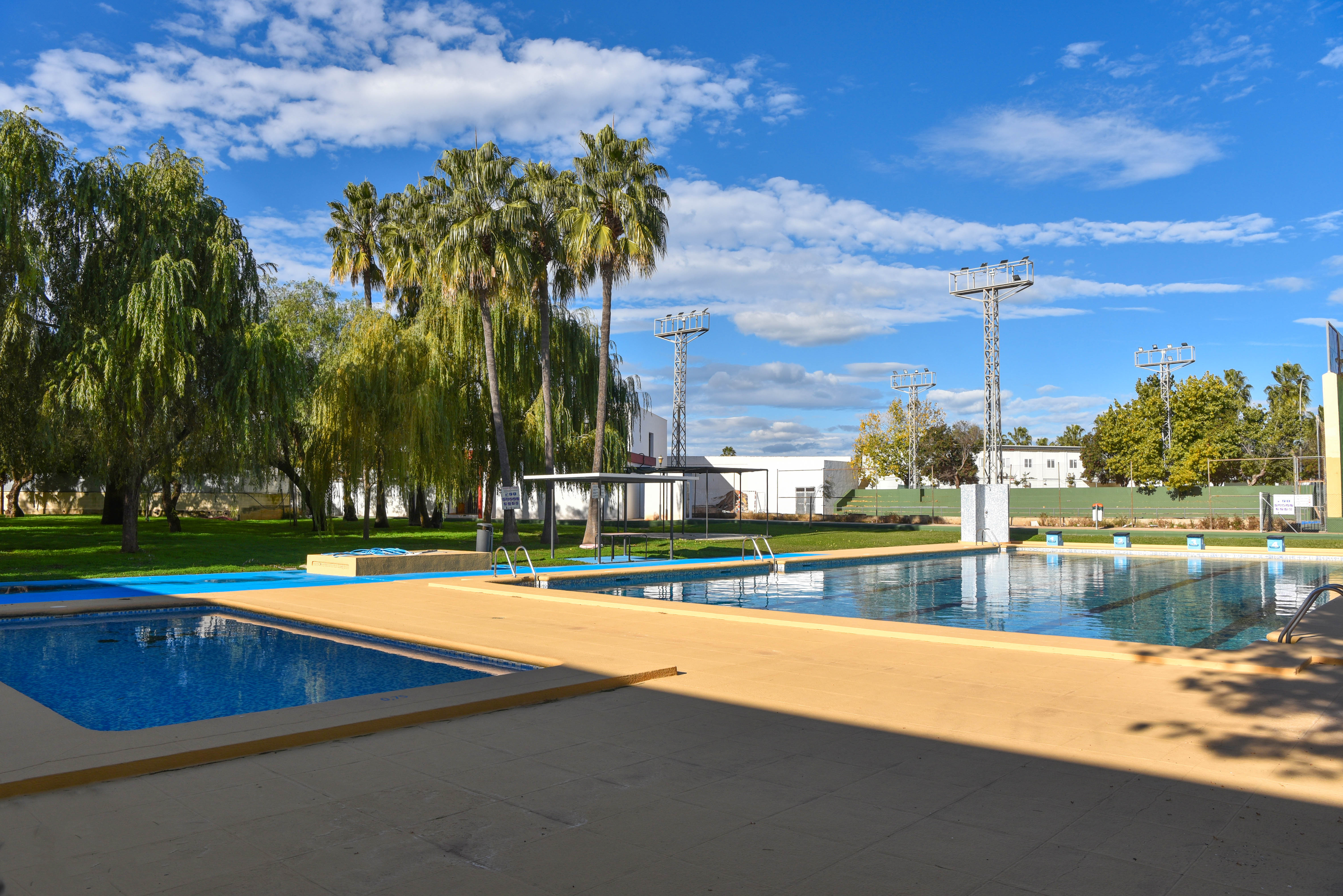 Municipal swimming pool complex featuring a main pool, children's pool, sun deck, and lawn area with mature palms and sports lighting.