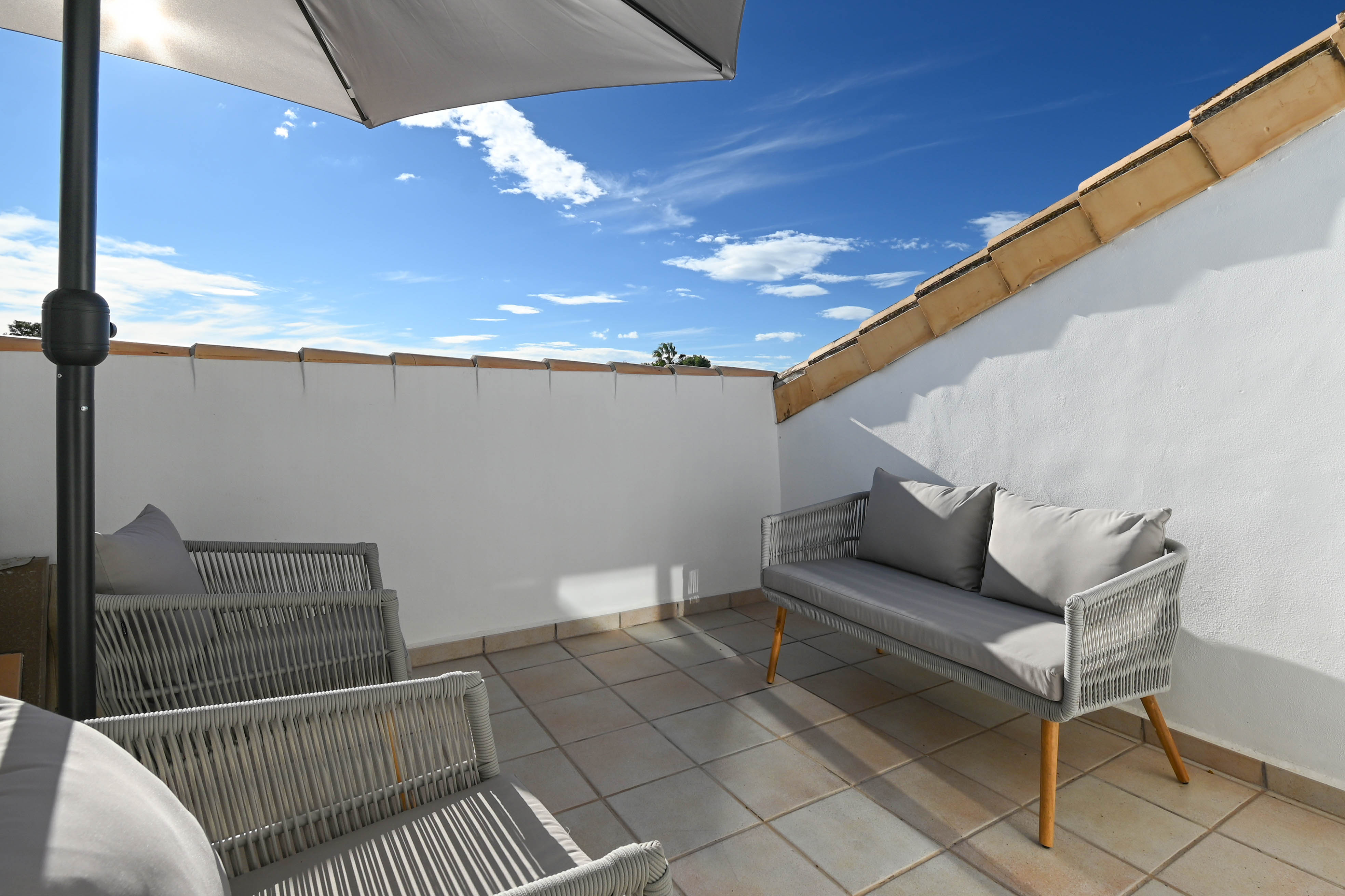 Private rooftop terrace featuring grey woven outdoor seating, tiled flooring, and a parasol, set against white walls and terracotta roof tiles.