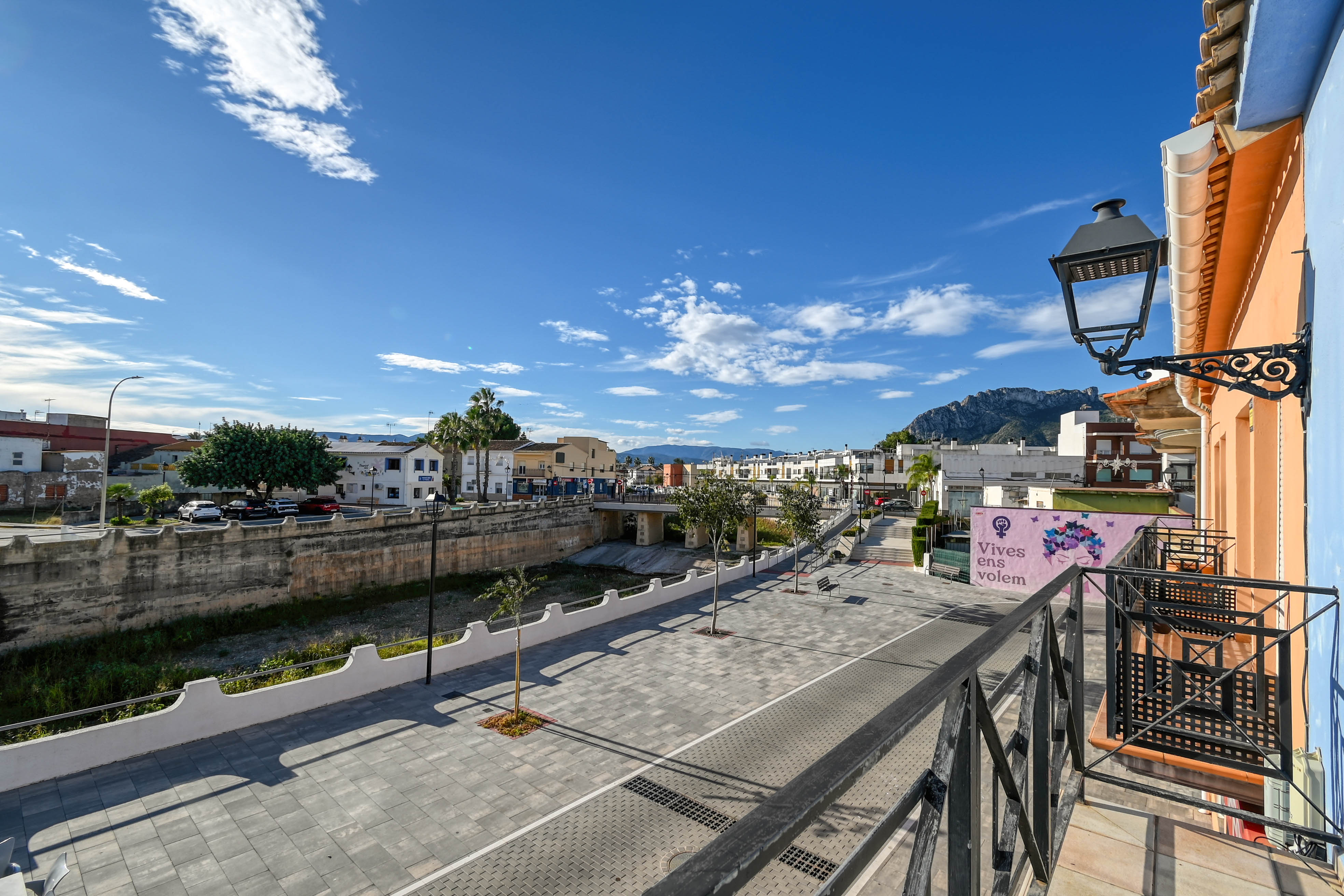 View from the balcony with black iron railings, overlooking a paved pedestrian plaza, the stone-walled riverbed, and distant mountain peaks.
