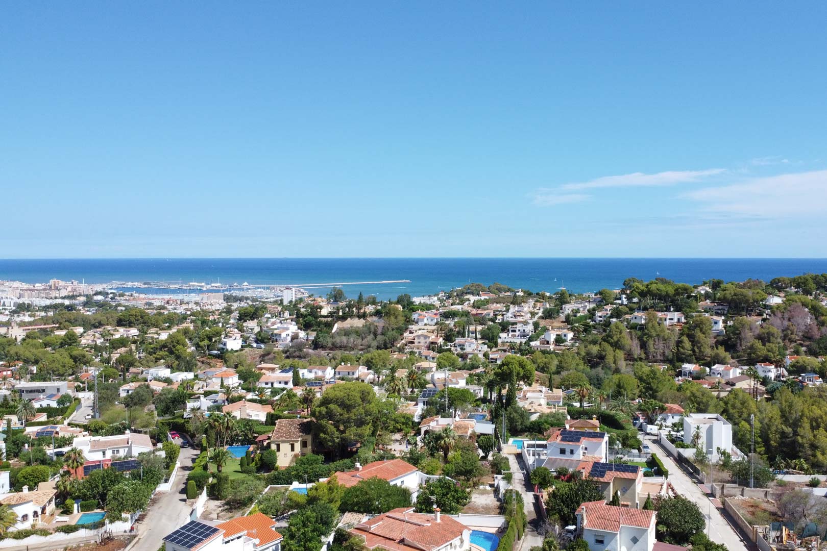 Aerial perspective of the Dénia residential district overlooking the Mediterranean Sea and the local port, featuring villas with pools and lush Mediterranean vegetation.