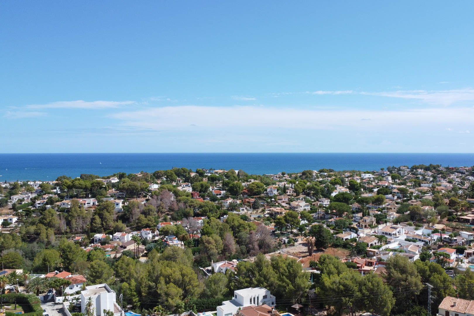 Panoramic aerial view of a residential neighborhood in Dénia, featuring white villas set within lush pine groves and a clear view of the Mediterranean Sea.
