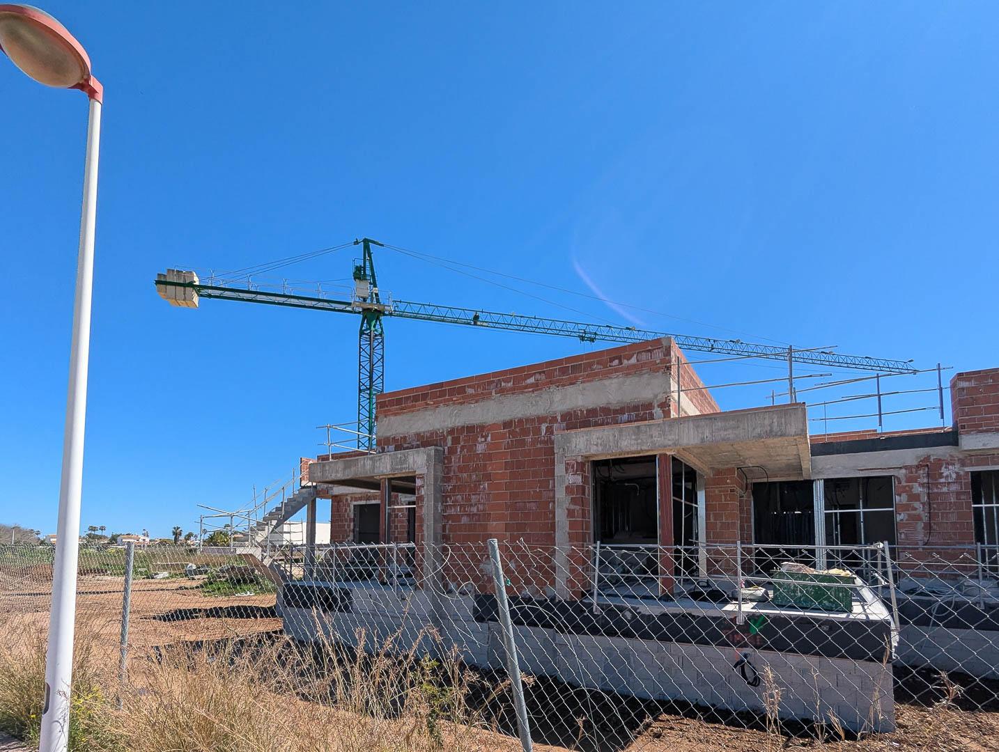 Current construction phase showing the brick and concrete shell of the villa, featuring an active crane and perimeter fencing under a clear blue sky.