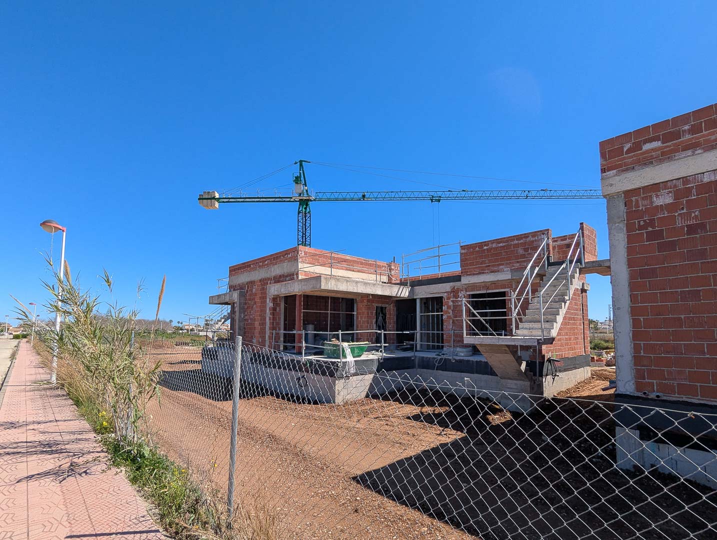 Current construction progress featuring the red brick structure, concrete slabs, and external staircase on a flat plot with a crane in the background.