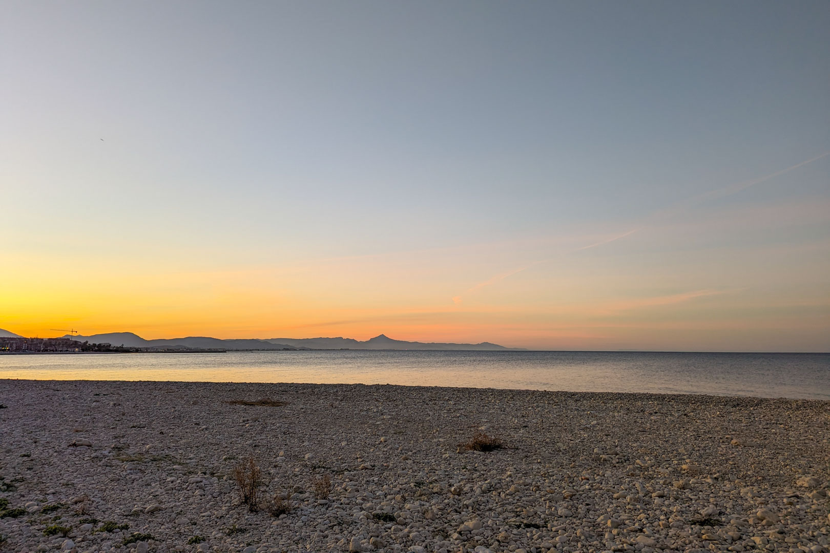 Panoramic view of the pebble beach near the property at sunset, featuring distant mountain silhouettes and calm Mediterranean waters.