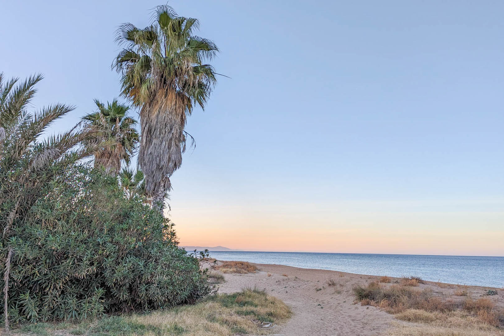 View of the nearby beach featuring sandy dunes, palm trees, and Mediterranean shrubs under a soft twilight sky, located less than two kilometers from the property.