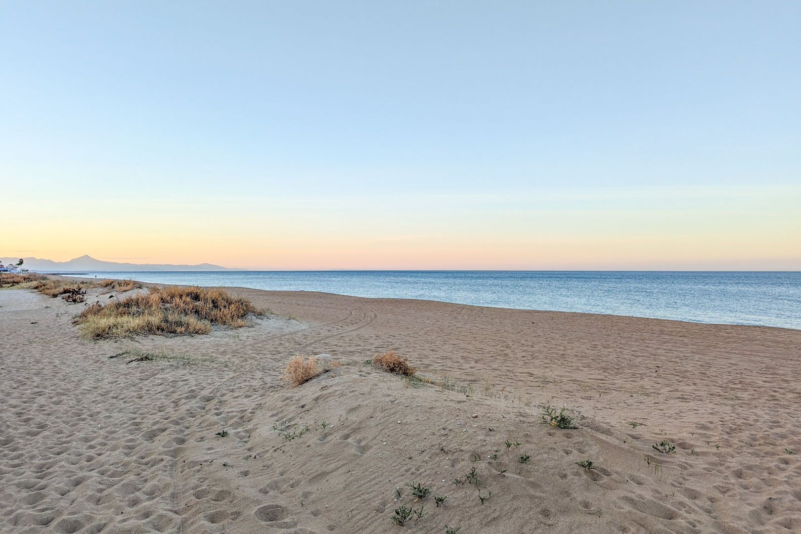Wide sandy beach with natural dunes and native vegetation located near the property. Features calm Mediterranean waters and distant mountain silhouettes at dusk.