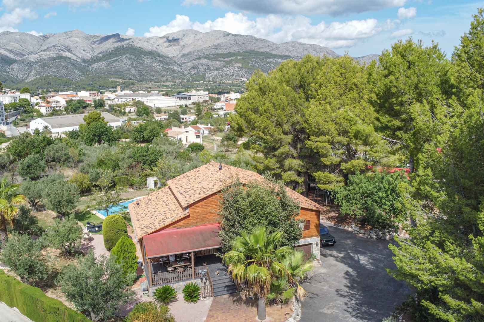 Aerial view of the Finnish pine villa featuring a tiled roof, surrounded by mature trees and gardens with views of the Orba valley and mountains.