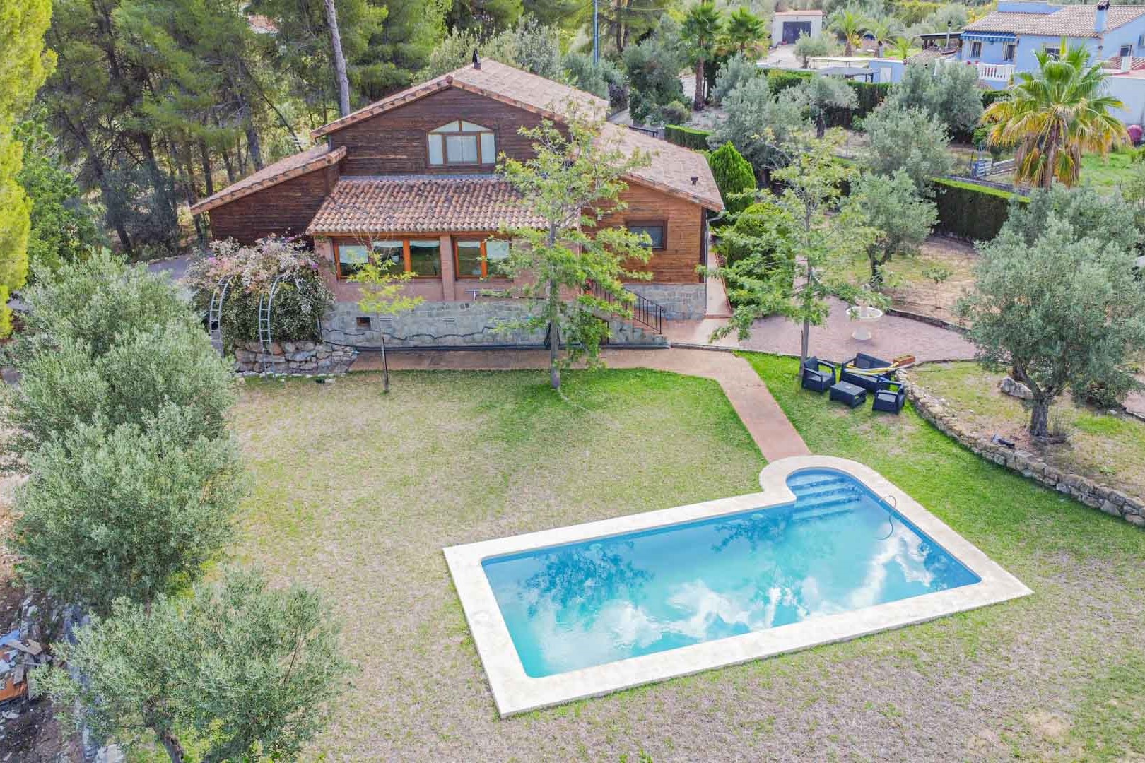 Aerial view of the villa featuring wood and stone architecture, a private swimming pool with integrated steps, and a lawn bordered by olive trees.