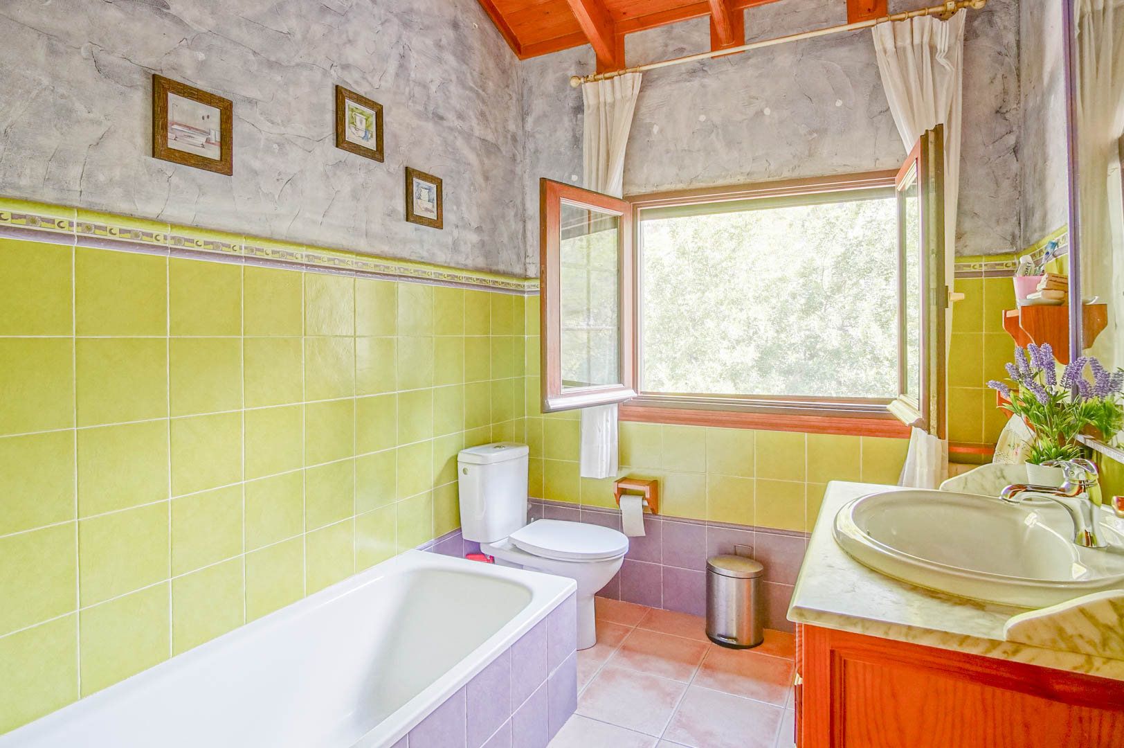 Bathroom featuring exposed wooden ceiling beams, green tiling, and a marble-top vanity. A large window provides natural light and views of the surrounding greenery.