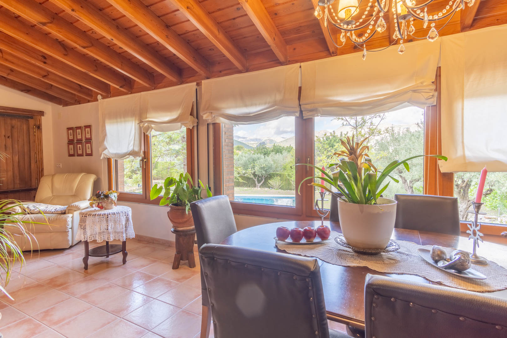 Bright dining area featuring exposed Finnish pine ceiling beams, terracotta flooring, and large windows overlooking the private pool and Mediterranean landscape.