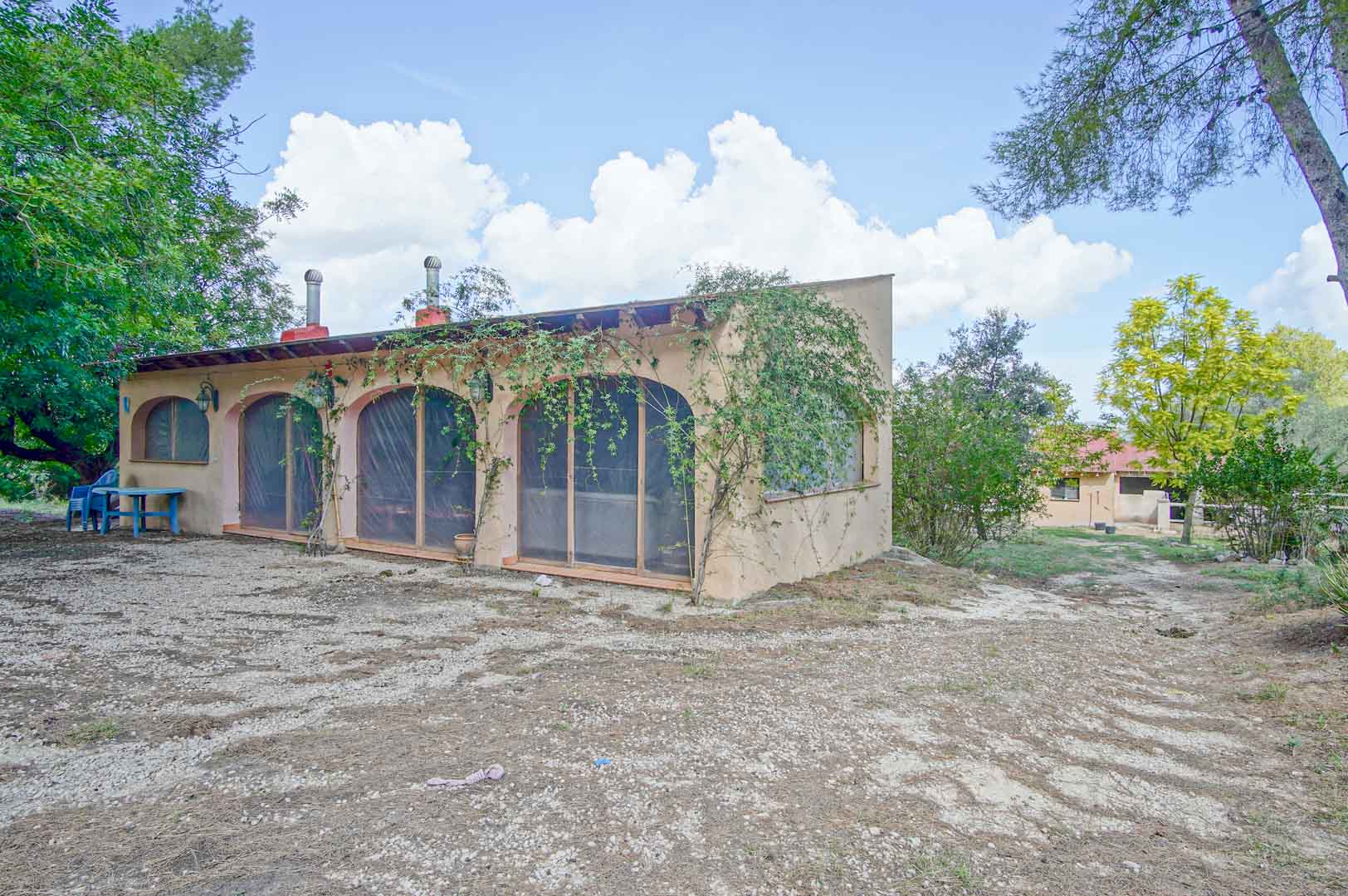 Exterior outbuilding featuring arched openings with screens and exposed wooden beams, set within a natural landscape of pines and Mediterranean flora.