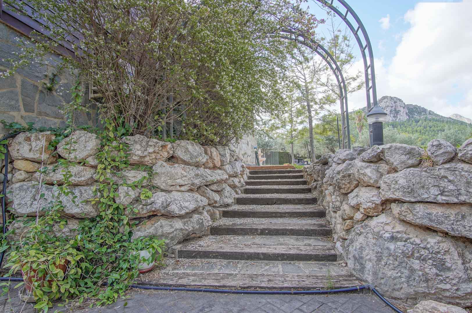 Exterior staircase featuring wooden sleepers and natural stone walls, framed by a metal archway with views of the surrounding mountain landscape.