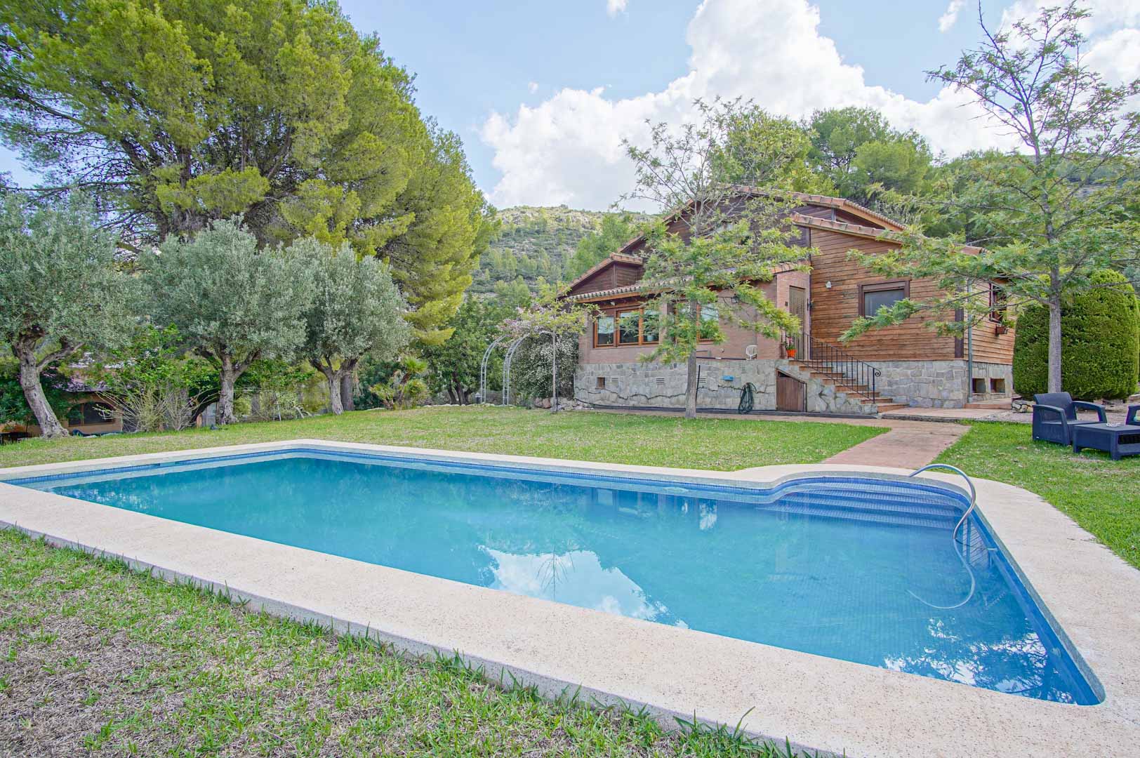 Exterior view featuring a rectangular swimming pool and the villa's facade, constructed with Finnish pine and natural stone, set against a backdrop of Mediterranean pines and mountains.