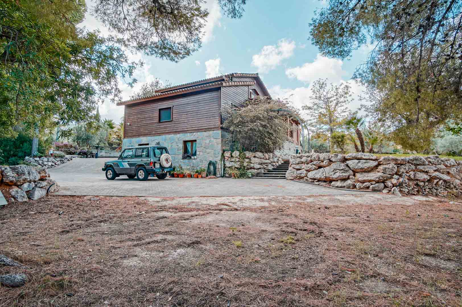 Exterior view of the villa featuring natural stone and Finnish pine cladding, a spacious paved driveway, and traditional stone retaining walls.