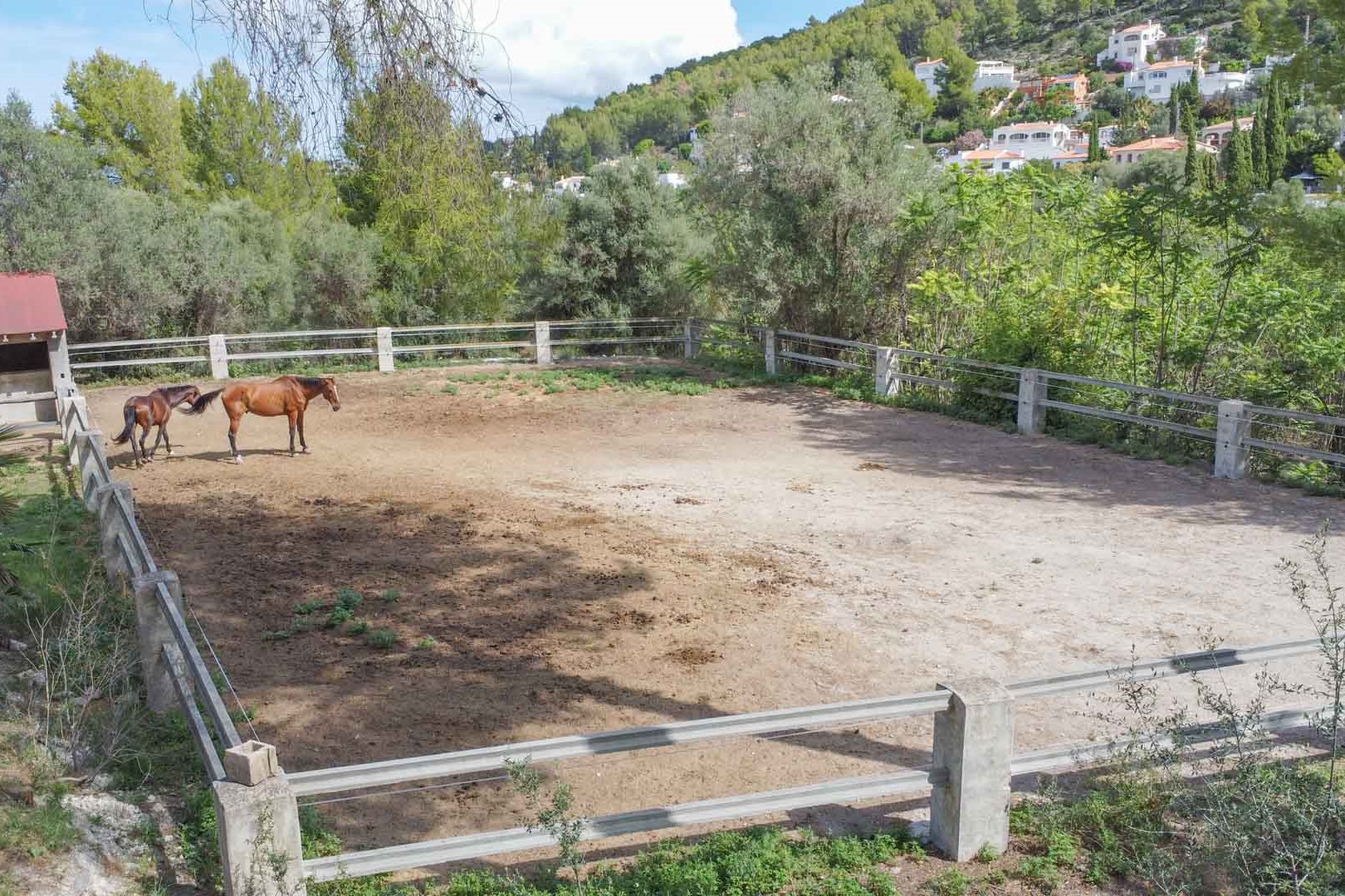 Fenced equestrian paddock featuring stone pillars and horizontal rails, set within a vast Mediterranean landscape with hillside views.