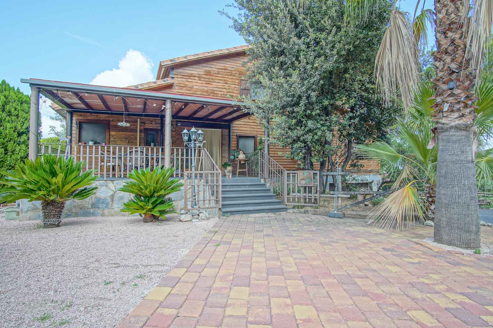 Finnish pine wood exterior featuring an elevated porch with a stone base, metal railings, and a paved entrance surrounded by Mediterranean palms and gravel.