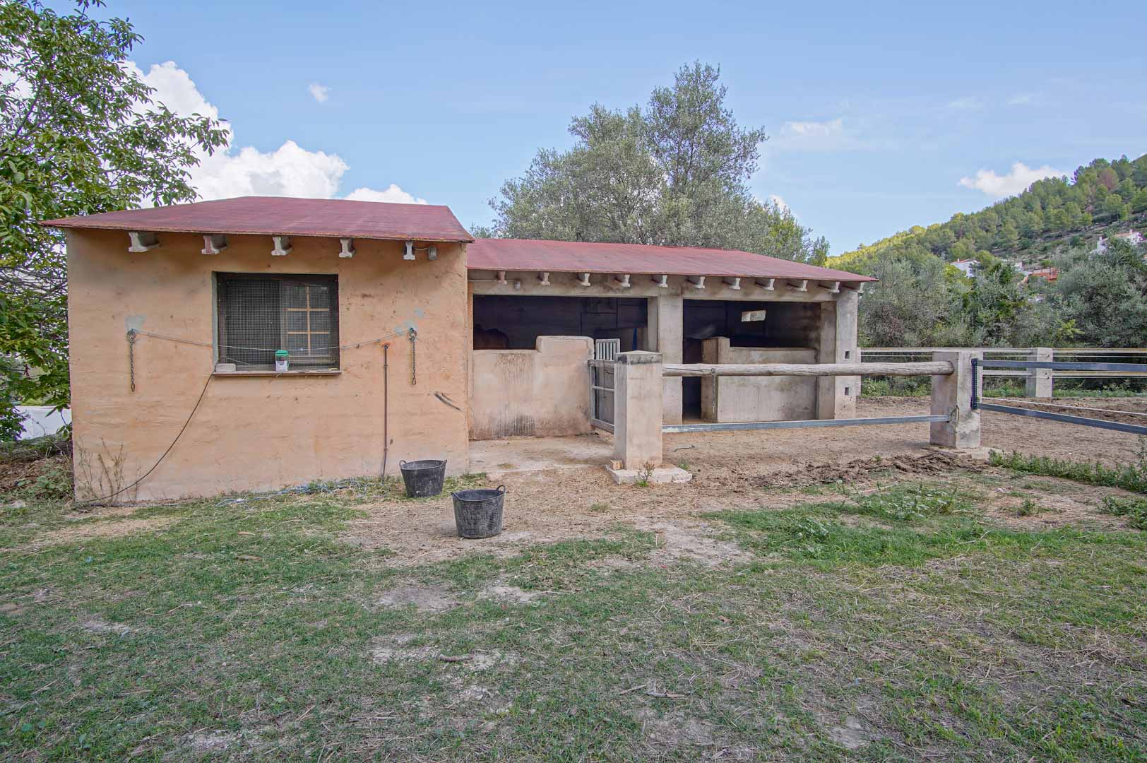 Functional horse stables featuring open stalls and a secure paddock with wood and metal fencing, set against a backdrop of Mediterranean hills.