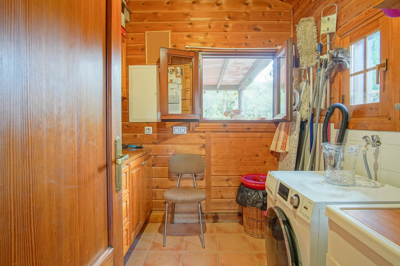 Functional laundry room with Finnish pine walls, featuring a washing machine, storage cabinetry, and a window overlooking the garden.