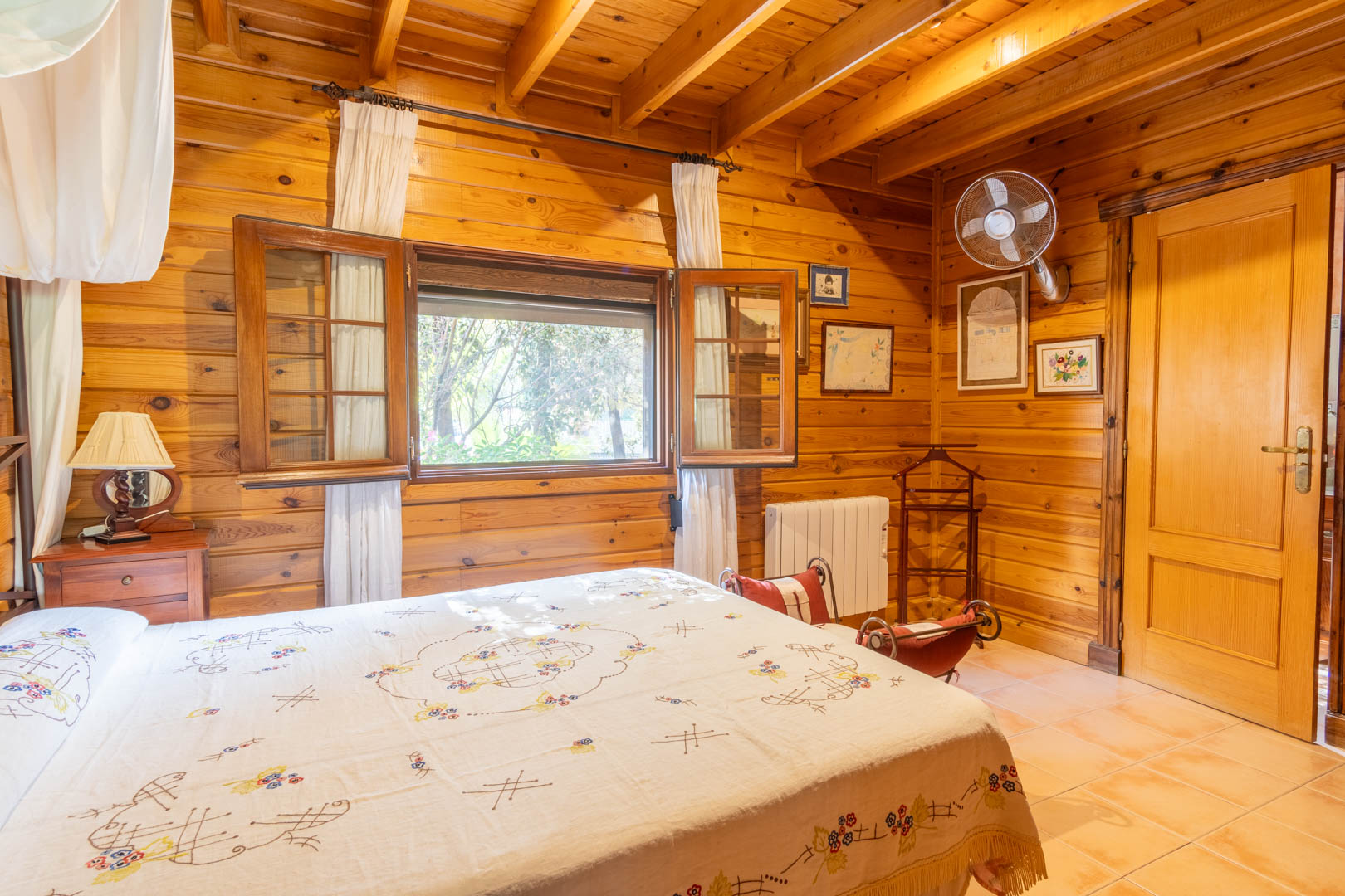 Rustic bedroom featuring full Finnish pine wood paneling, exposed beam ceilings, ceramic tile flooring, and a wooden window with shutters providing natural light.