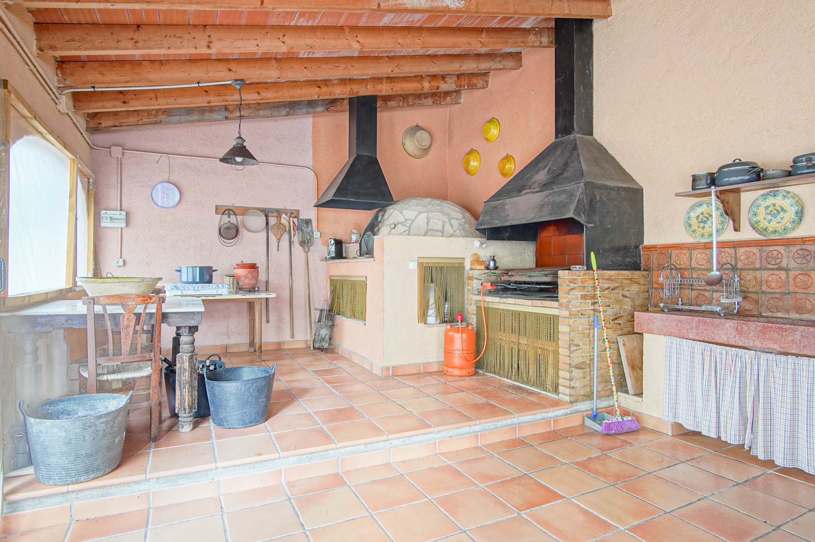 Rustic outdoor kitchen featuring a traditional wood-fired stone oven, brick barbecue with metal hood, and exposed timber beams.