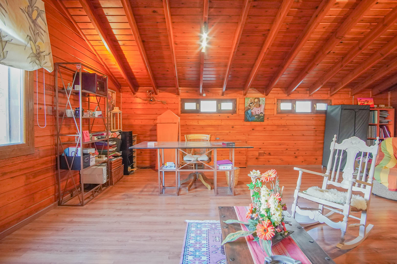 Spacious attic office featuring Finnish pine walls and exposed ceiling beams. The layout includes a glass-top desk and a dedicated reading area with a classic white rocking chair.