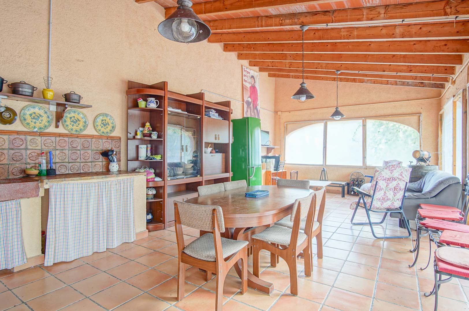 Spacious rustic gathering area featuring exposed wooden beams, terracotta flooring, an integrated kitchen, and a dining space with large windows.