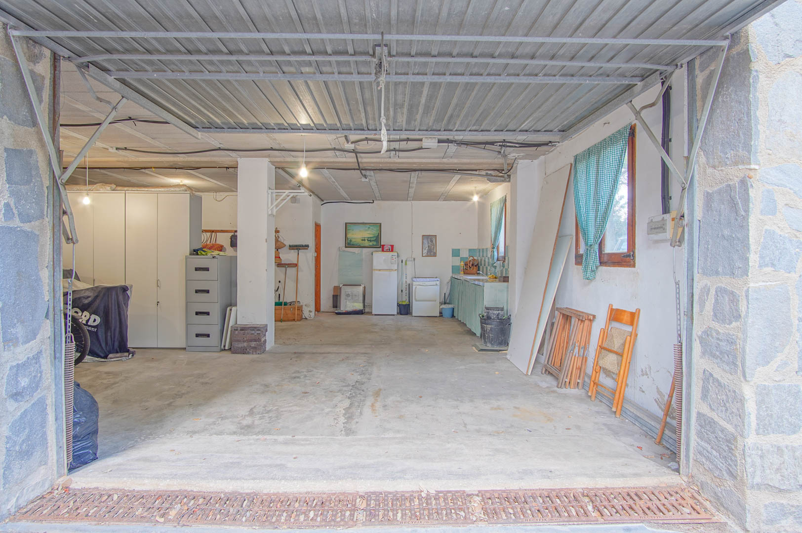 Spacious semi-basement featuring a multi-car garage, workshop area, and integrated kitchenette. It includes concrete flooring, stone pillars, and a functional metal overhead door.