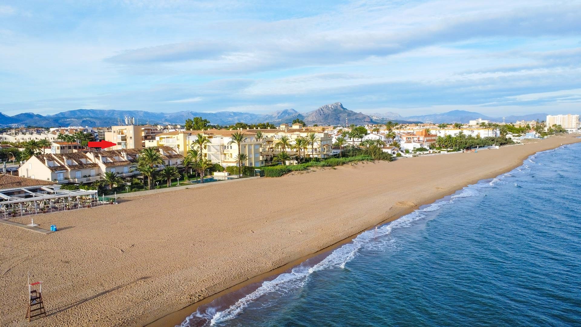Aerial perspective of the wide sandy beach and Mediterranean coastline, highlighting the apartment's location relative to the sea and mountains.