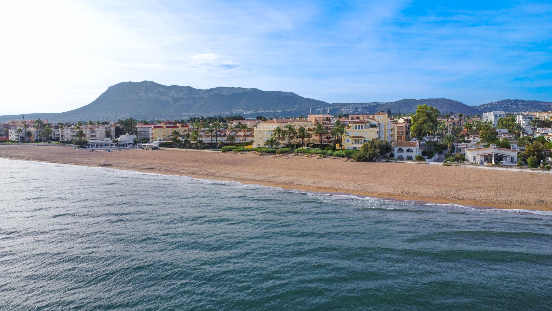 Aerial view of a sandy beachfront with residential complexes and palm trees, set against a backdrop of a prominent mountain range under a clear sky.