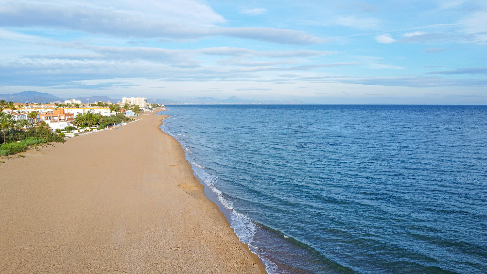Aerial view of a wide sandy beach bordering the Mediterranean Sea, featuring low-rise coastal residences and distant mountain silhouettes under a clear sky.