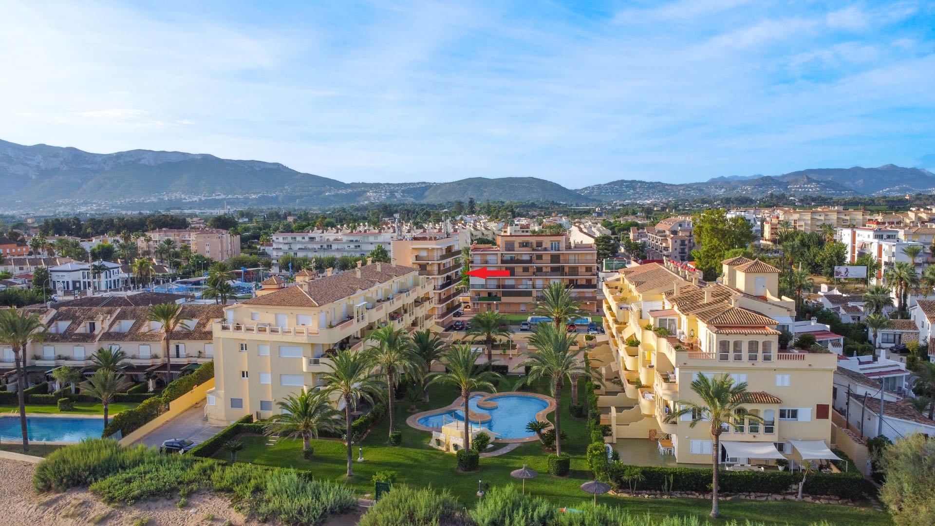 Aerial view of the residential complex showing the communal pool, landscaped gardens, and the specific unit location highlighted against a mountain backdrop.