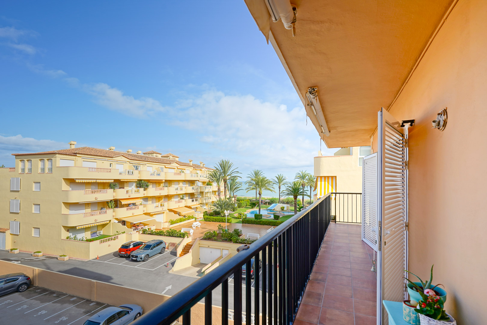 Balcony with terracotta tiles and black railing offering views of the communal pool, landscaped gardens, and the Mediterranean horizon.