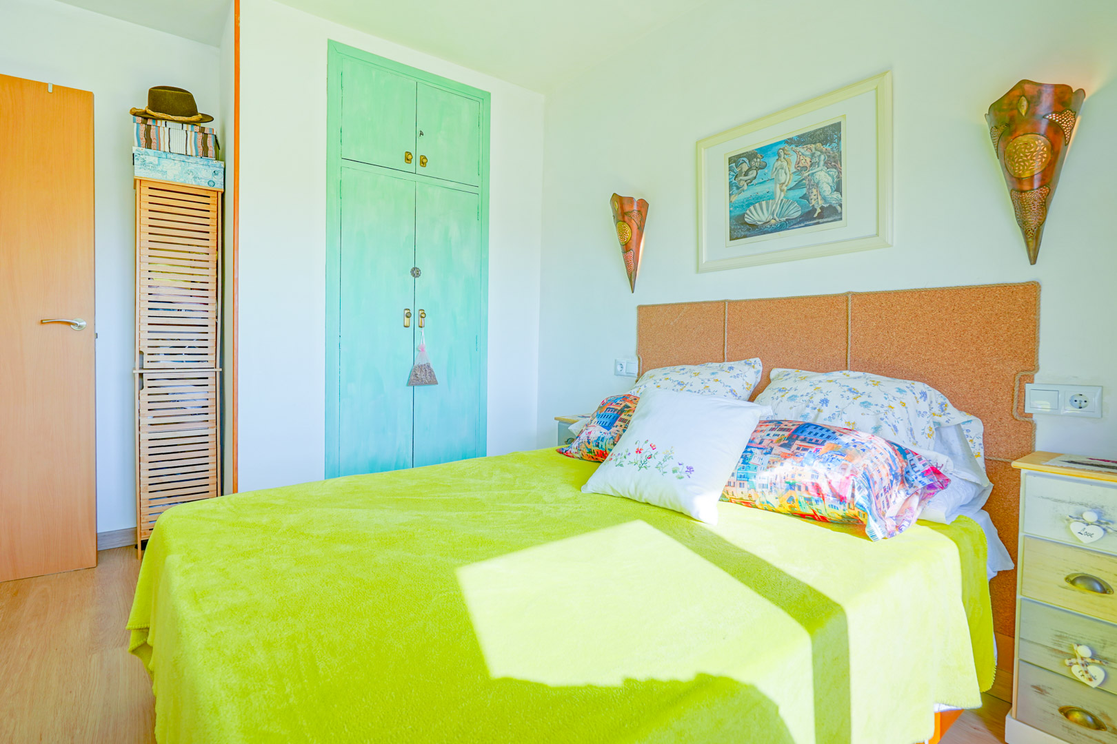 Bedroom featuring a cork headboard, mint green built-in wardrobe, and Botticelli artwork. Natural light illuminates the lime green bedding and rustic bedside table.