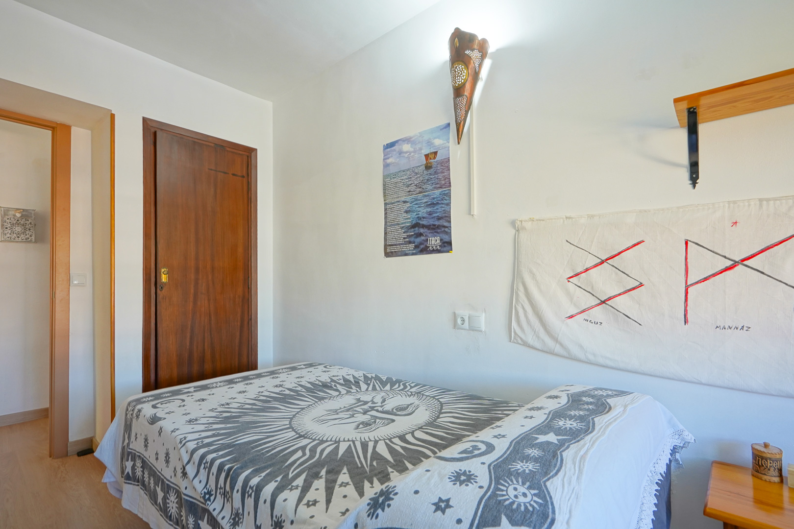 Bedroom featuring a single bed with a sun-motif bedspread, white walls decorated with a copper-style wall sconce, and a solid dark wood door.