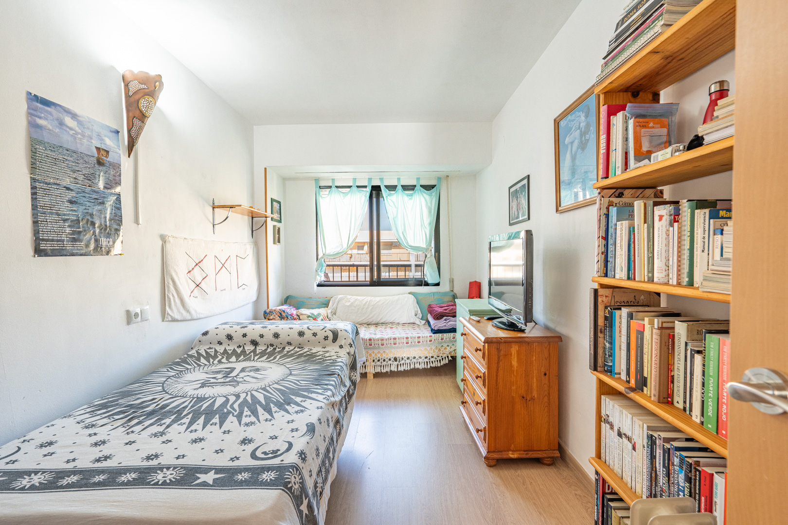 Bedroom with a single bed, wooden shelving, and a dresser. Features laminate flooring and a window with light blue curtains providing natural light.