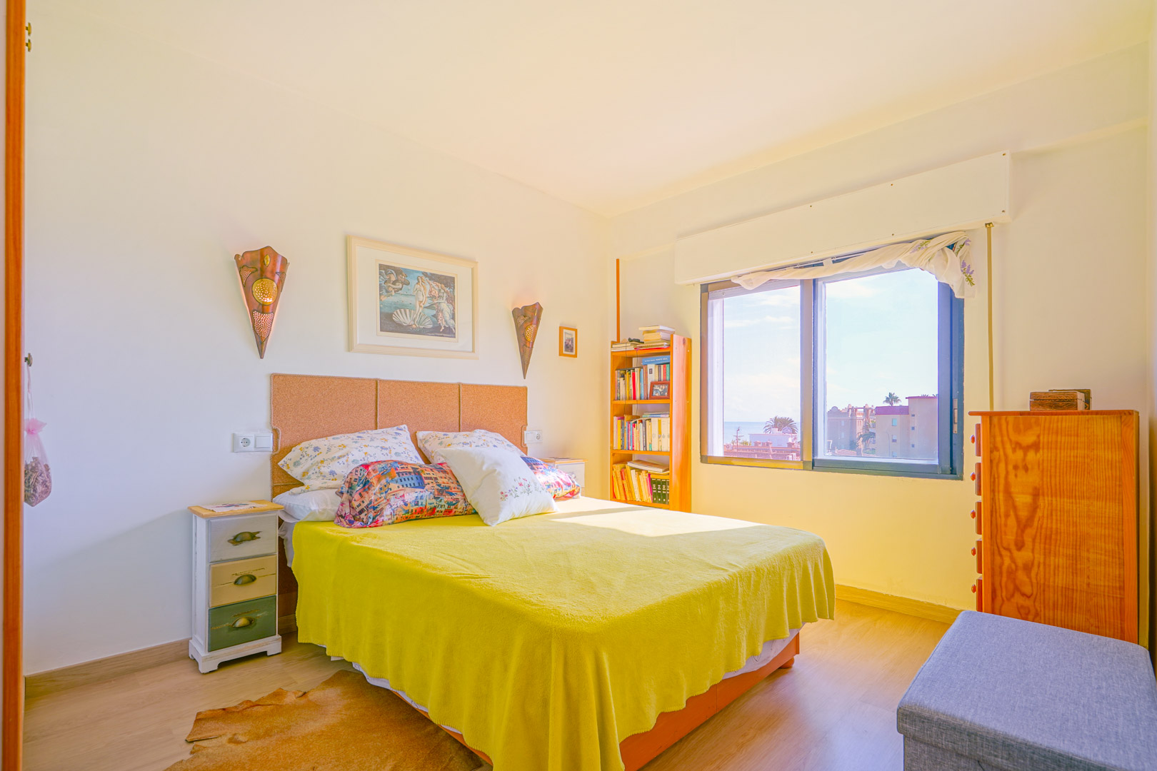 Bright bedroom featuring a cork headboard, yellow bedspread, and wooden furniture. A large window provides natural light and views of the neighborhood.