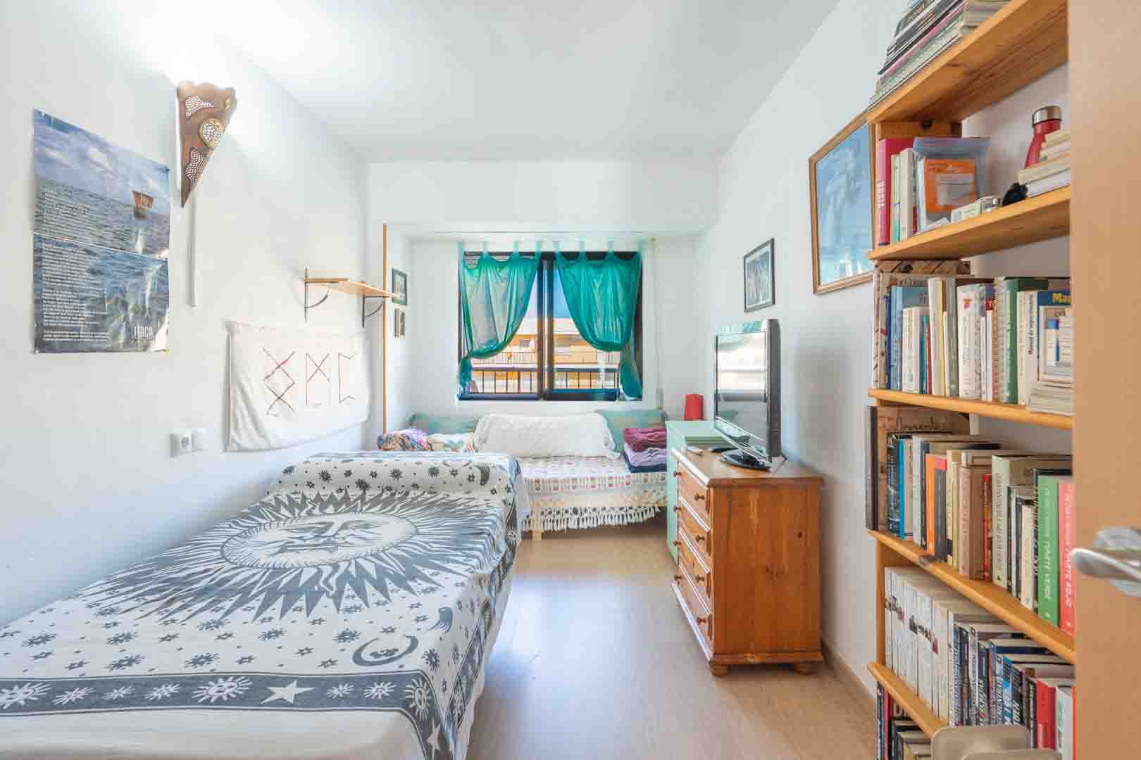Bright bedroom featuring a single bed with celestial patterns, a wooden bookshelf, and a dresser with a TV. Natural light enters through a window with turquoise curtains.