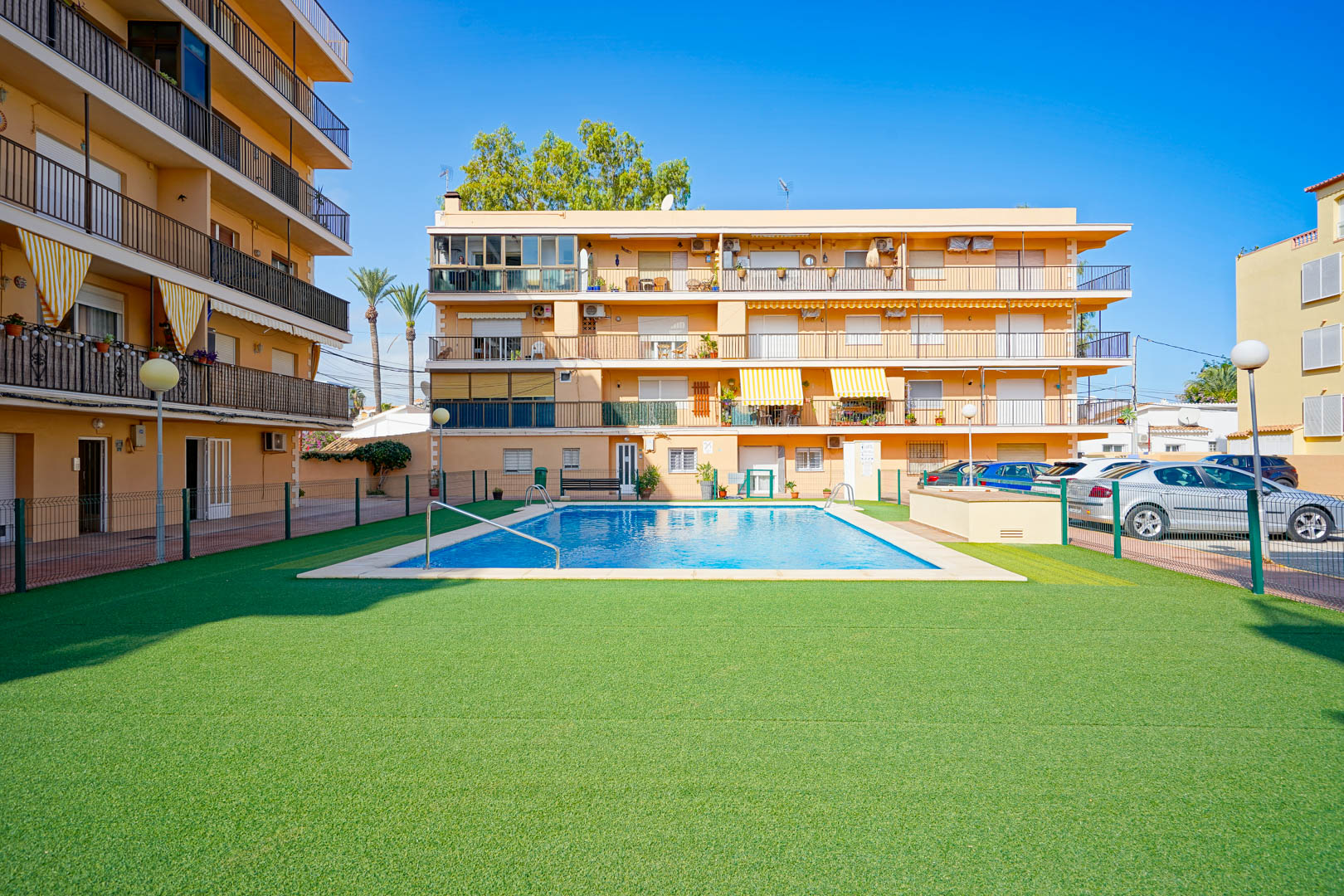Communal rectangular swimming pool surrounded by a spacious artificial grass area and a secure perimeter fence within a residential complex.