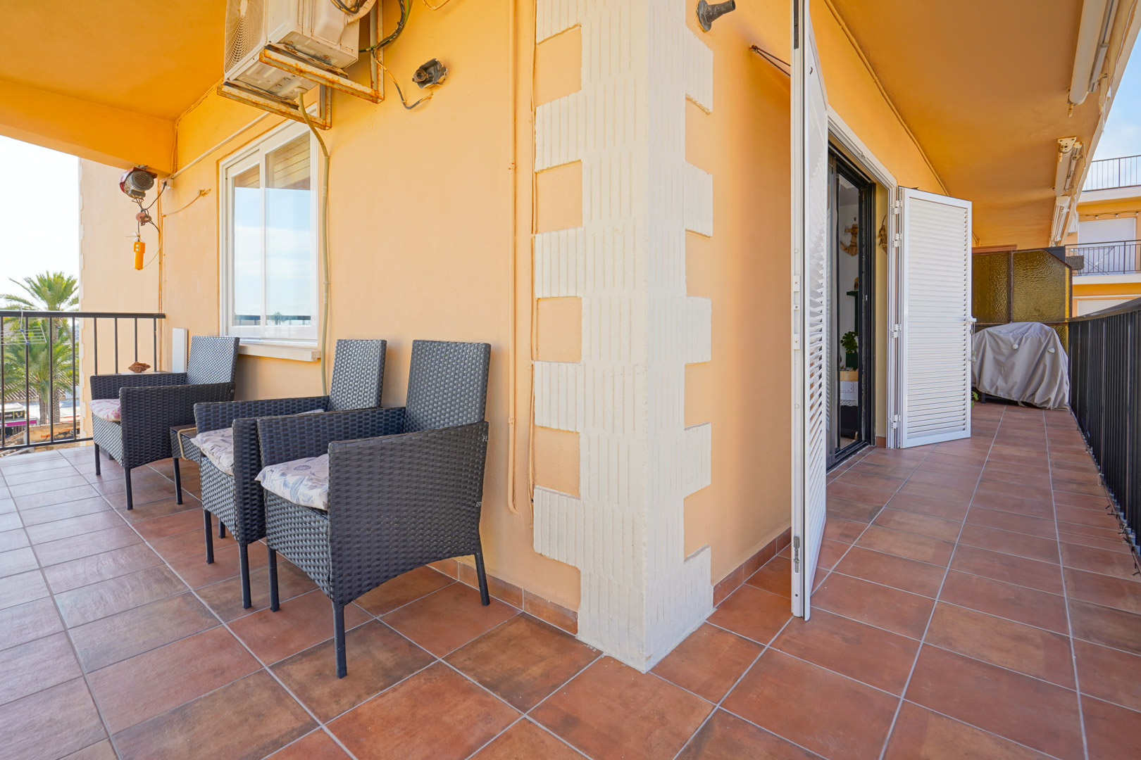 Covered terrace with terracotta floor tiles, dark wicker armchairs, and white shutters. The space includes a wall-mounted air conditioning unit and metal railings.