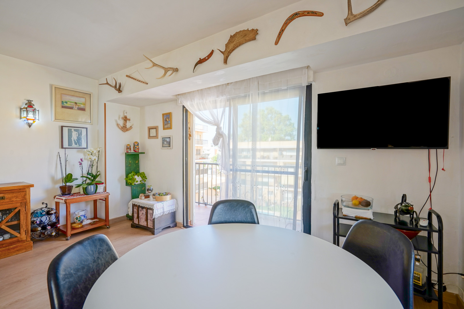 Dining area with a white circular table, wall-mounted TV, and balcony access. Unique wall decor includes mounted antlers and boomerangs above the sliding door.