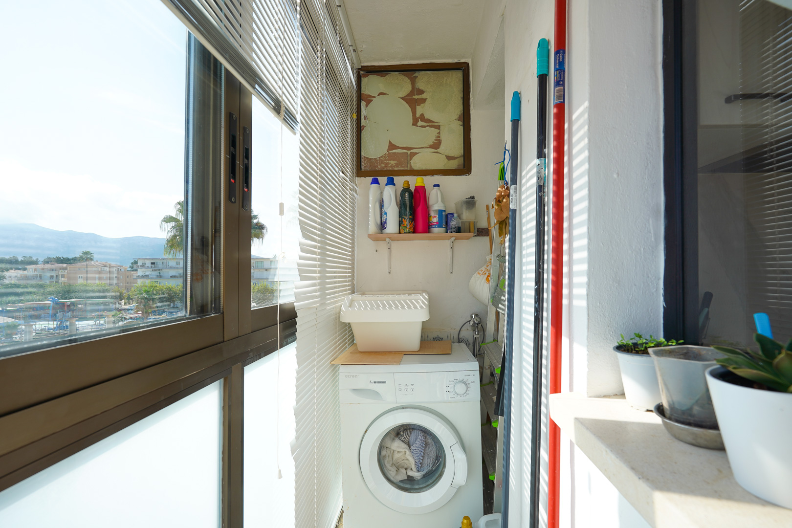 Enclosed utility balcony featuring a front-loading washing machine, storage shelf for cleaning supplies, and large windows with horizontal blinds.